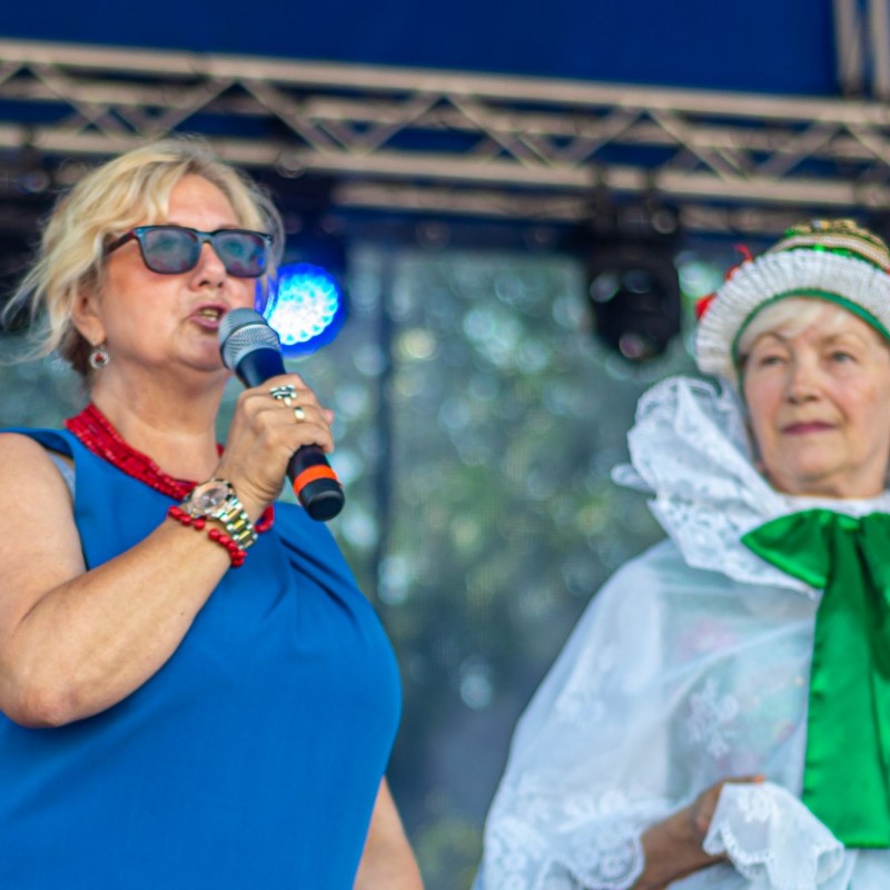 Fot. Łukasz Kuc/Na scenie plenerowej kobieta zapowiada przez mikrofon, obok niej stoi kobieta ubrana w strój.