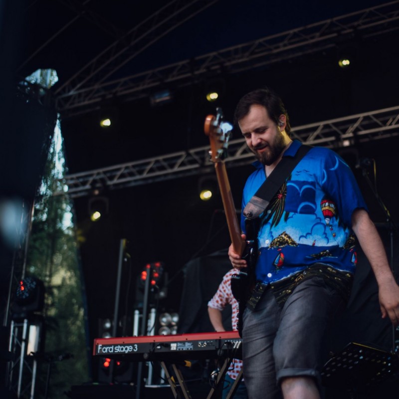 Fot. Łukasz Kuc/Członek zespołu muzycznego w kolorowej letniej koszuli na scenie plenerowej, w tle pianista.