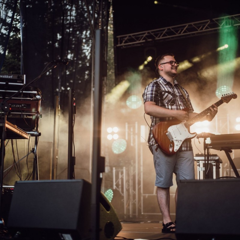 Fot. Łukasz Kuc/Uśmiechnięty gitarzysta na scenie plenerowej, w tle światła reflektorów.