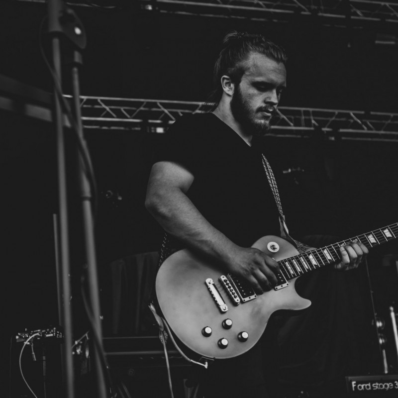 Fot. Łukasz Kuc/Czarno-białe zdjęcie gitarzysty w białek koszulce występującego na scenie plenerowej.