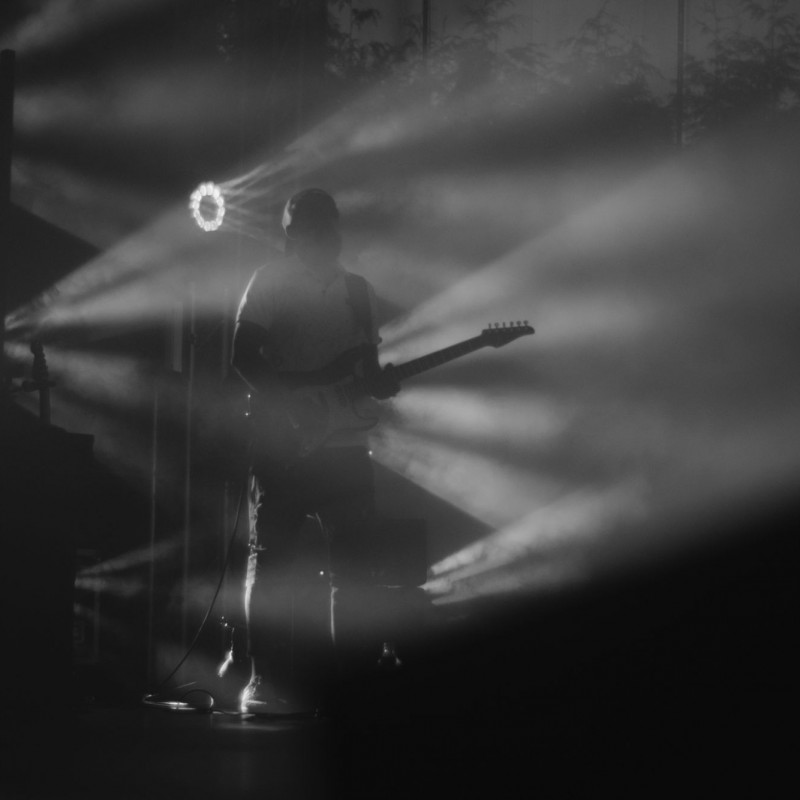 Fot. Łukasz Kuc/Czarno-białe zdjęcie gitarzysty podczas występu w świetle reflektorów i kłębach dymu.