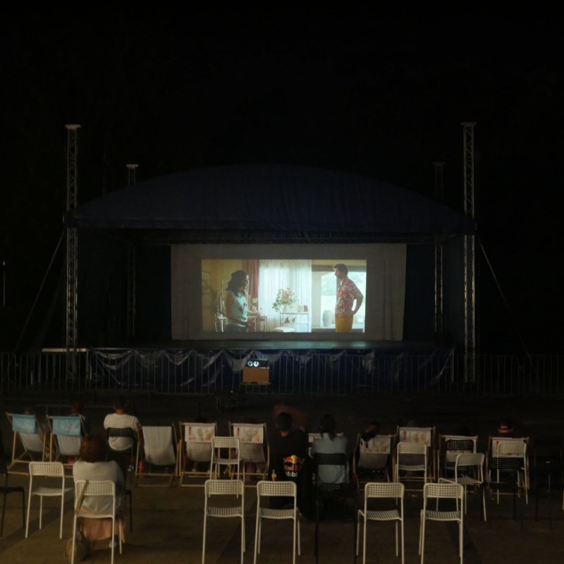 Ludzie siedzą na krzesłach i oglądają film w plenerze wyświetlany na dużym ekranie.