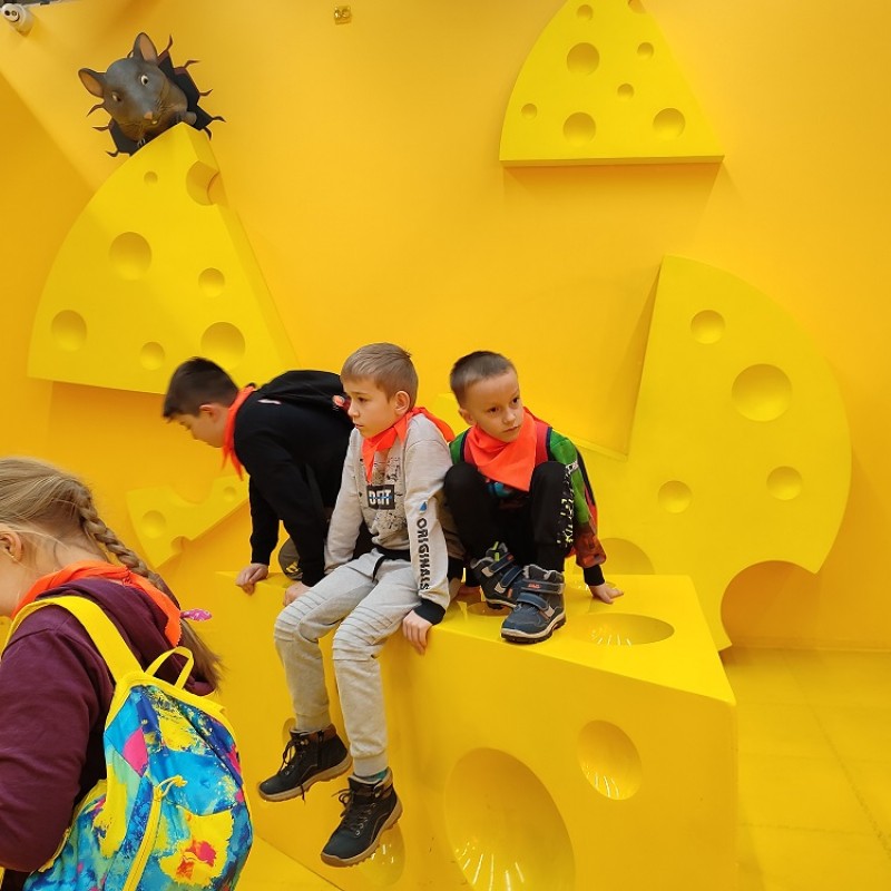 Dzieci siedzące na kostce sera żółtego z dziurami. W lewym górnym roku wychylająca się z norki makieta myszki.