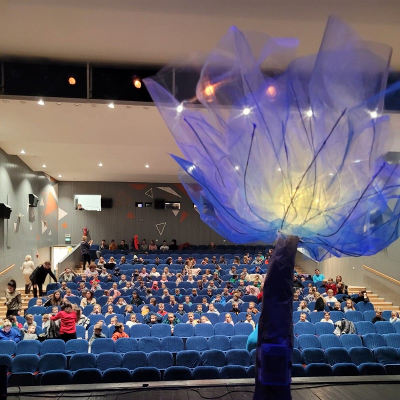 Ujęcie ze sceny DK na widzów w sali widowiskowej. Na pierwszym planie element scenografii przedstawiający duży, niebieski, oświetlony kwiat.